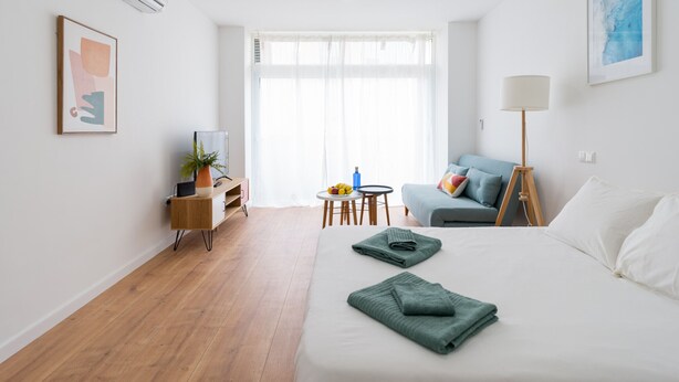 Quarto de hotel moderno com cama, escrivaninha, cadeira, luminária e janela com cortinas leves.