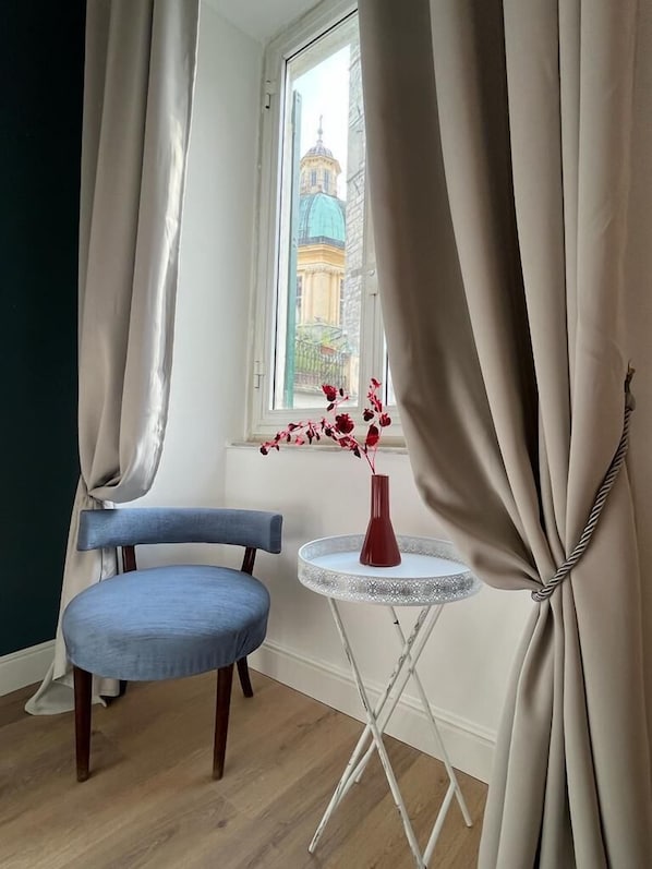 Um quarto com uma cadeira azul, uma mesa redonda pequena com um vaso de flores vermelhas e cortinas bege.