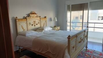 Uma cama grande com cabeceira decorada, um criado-mudo com abajur e vista da cidade através de grandes janelas.