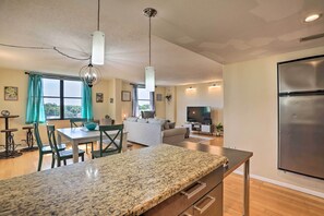 A modern kitchen with granite countertops, a dining area with a table and chairs, a living room with a sofa and TV, and a large window with curtains. - Convenient Clearwater Condo < 4 Mi to Beach! (Clearwater)