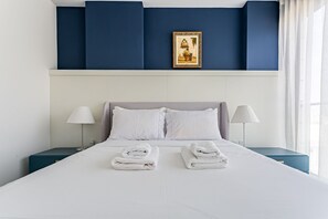 Uma cama bem arrumada com toalhas dobradas, um quadro emoldurado acima e duas mesas de cabeceira com abajures.