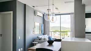 A modern living room with a grey sofa, a wooden dining table, and a large window with curtains. - WI-FI 300MB | Rooftop with Pool in Jurerê #JA15 (Florianópolis)
