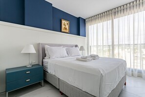Une chambre moderne avec un grand lit, deux tables de chevet et une vue sur la ville à travers de fines rideaux.