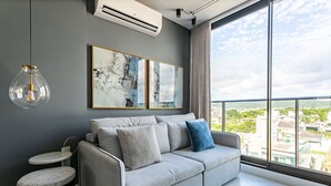 A modern living room with a grey sofa, two side tables, and a large window offering a city view. - WI-FI 300MB | Rooftop with Pool in Jurerê #JA15 (Florianópolis)