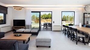 A modern living room with a flat-screen TV, a sofa, a dining area with a table and chairs, and large windows offering a view of the outdoors.