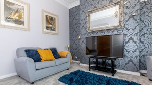 Ein Wohnzimmer mit grauem Sofa, blauen und gelben Kissen, einem großen Flachbildfernseher, einem blauen Teppich mit lockerem Flor und Wandtapete mit Muster.