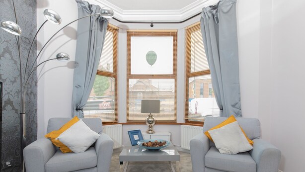 Ein Zimmer mit zwei grauen Sesseln, einem transparenten Glastisch, einer Lampe und einem Erkerfenster mit Vorhängen.