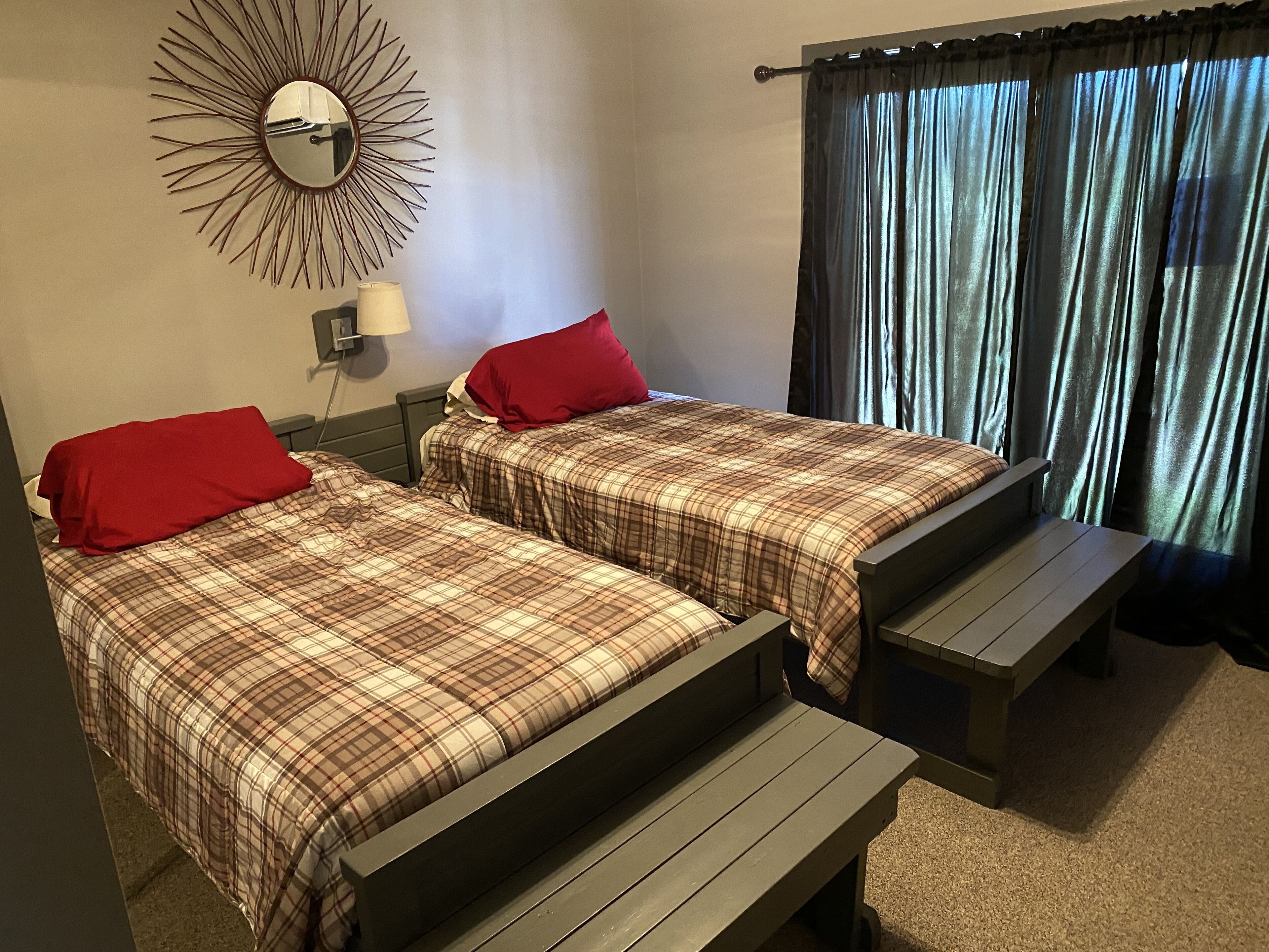 Two beds with plaid bedding and red pillows, a bench, a wall clock, and a window with curtains.