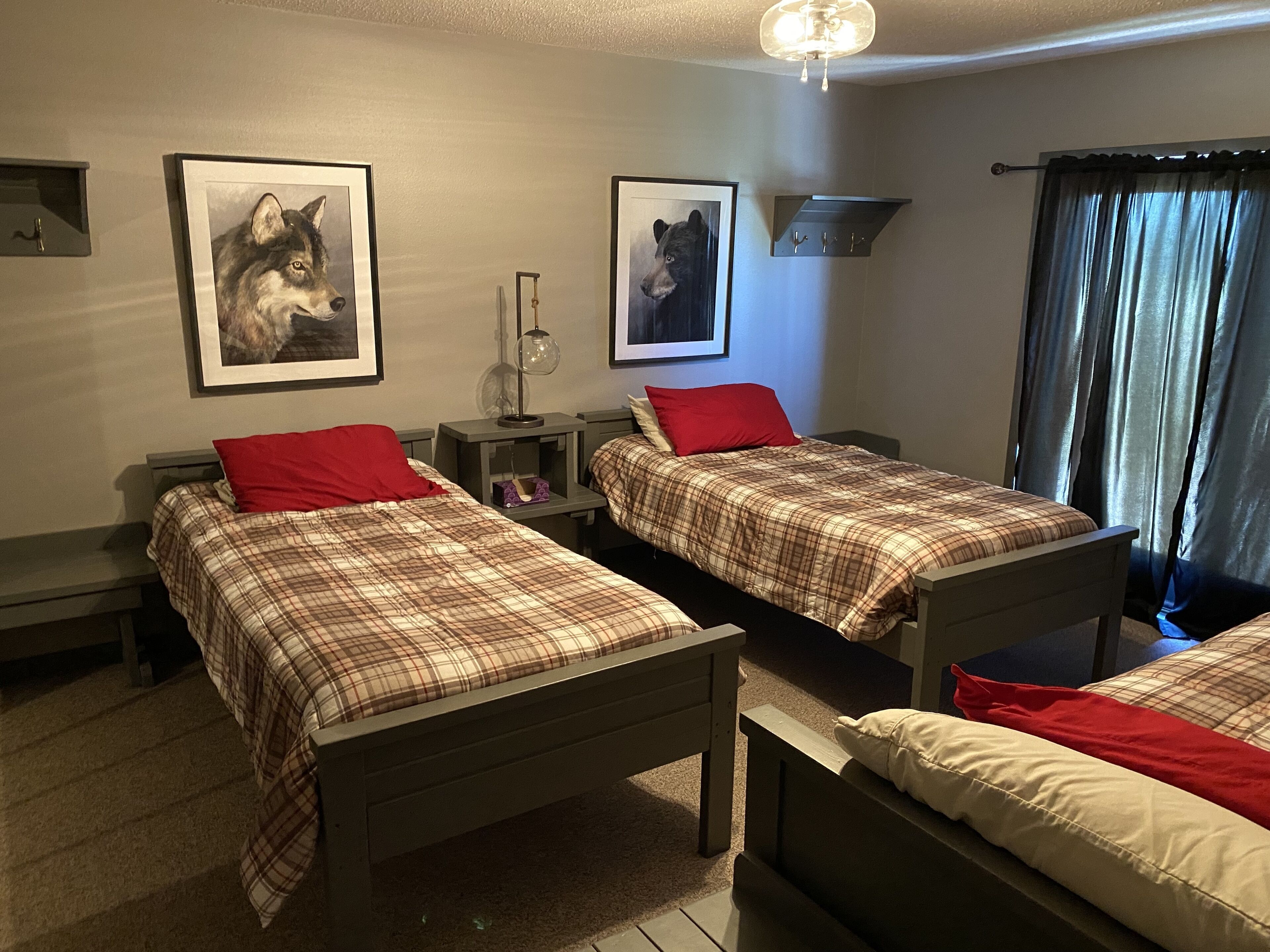 A room with two beds, each with plaid bedding and red pillows. There are framed animal pictures on the walls, a ceiling fan, and a window with curtains.