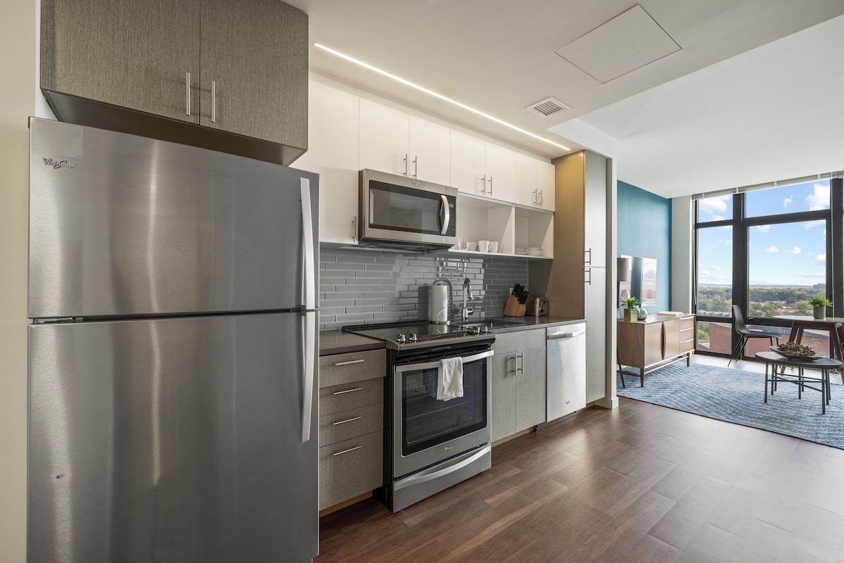A modern kitchen with stainless steel appliances, a microwave, and a stove. There is a dining area with a table and chairs, and a large window offering a view.