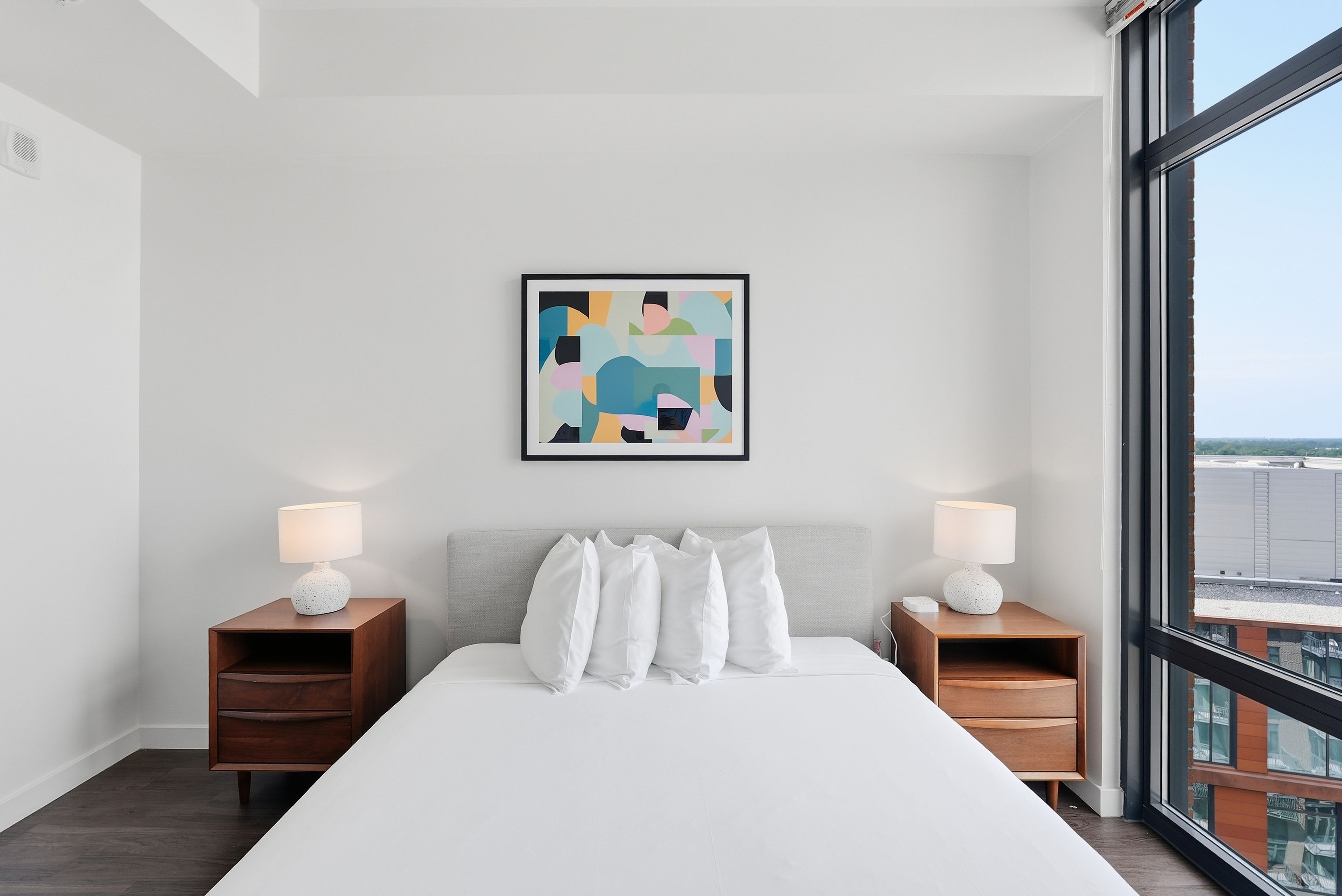 A modern hotel room with a large bed, two bedside tables with lamps, a framed abstract painting, and a large window offering a city view.