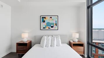 A modern hotel room with a large bed, two bedside tables with lamps, a framed abstract painting, and a large window offering a city view.