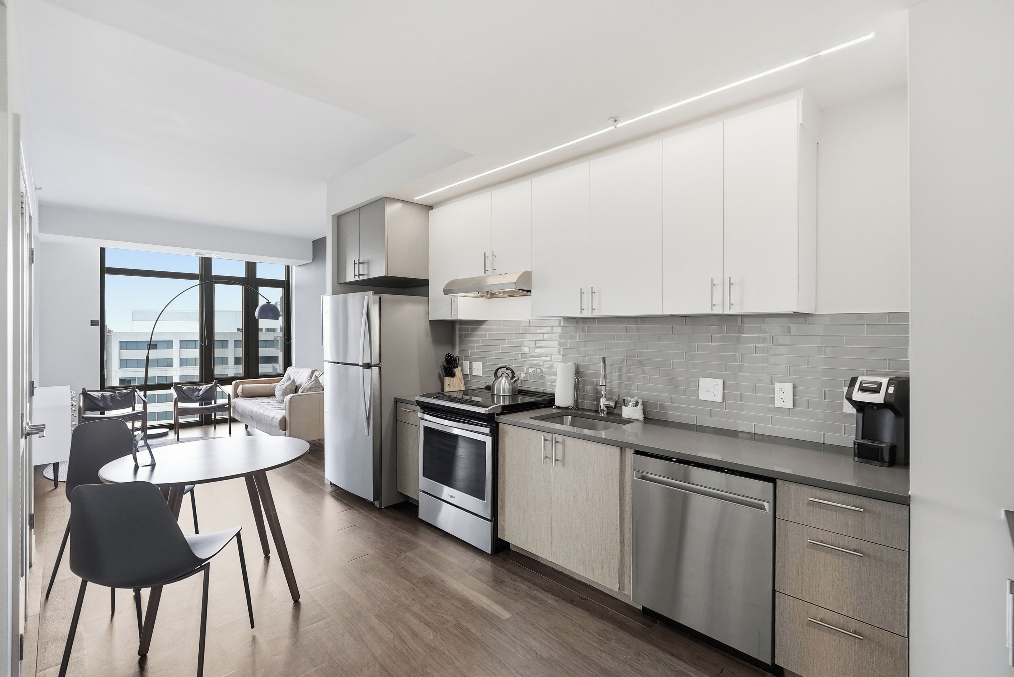 A modern kitchen with stainless steel appliances, a dining area with a round table and chairs, and a view of the city through large windows.