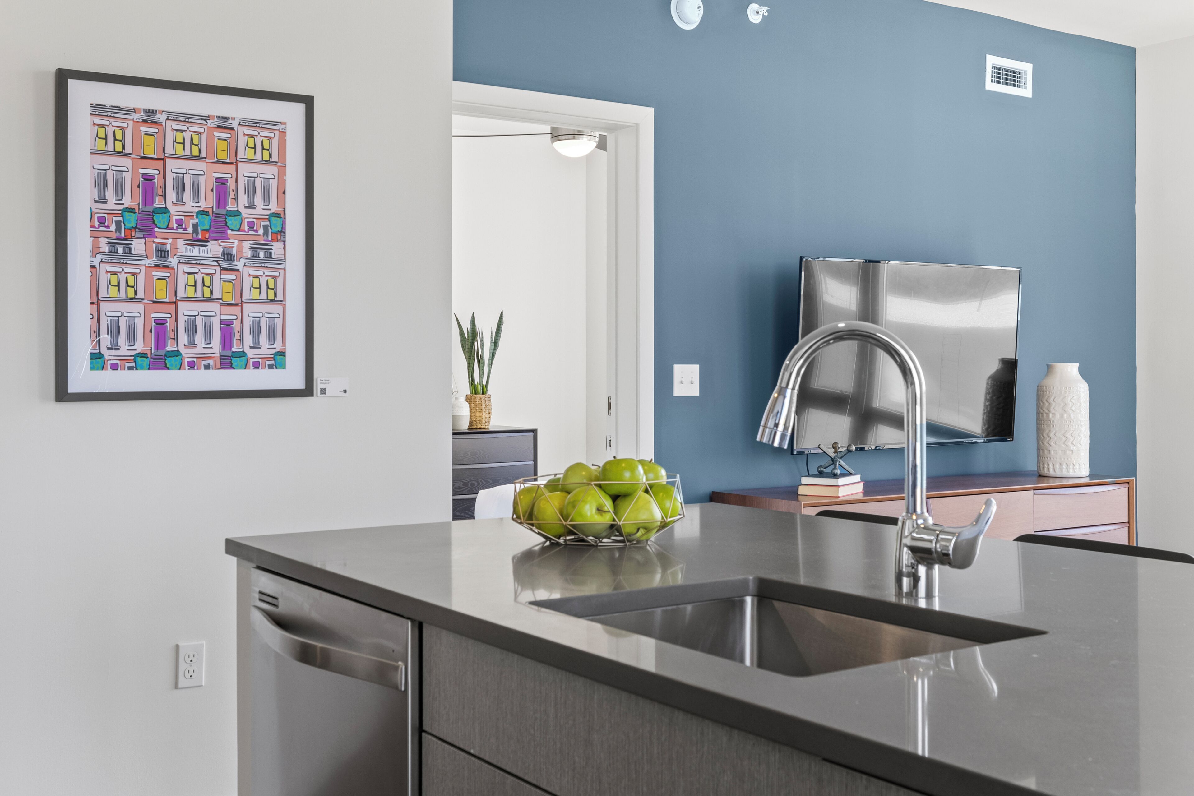 A modern kitchen with a stainless steel sink, a bowl of green apples, and a colorful artwork on the wall.