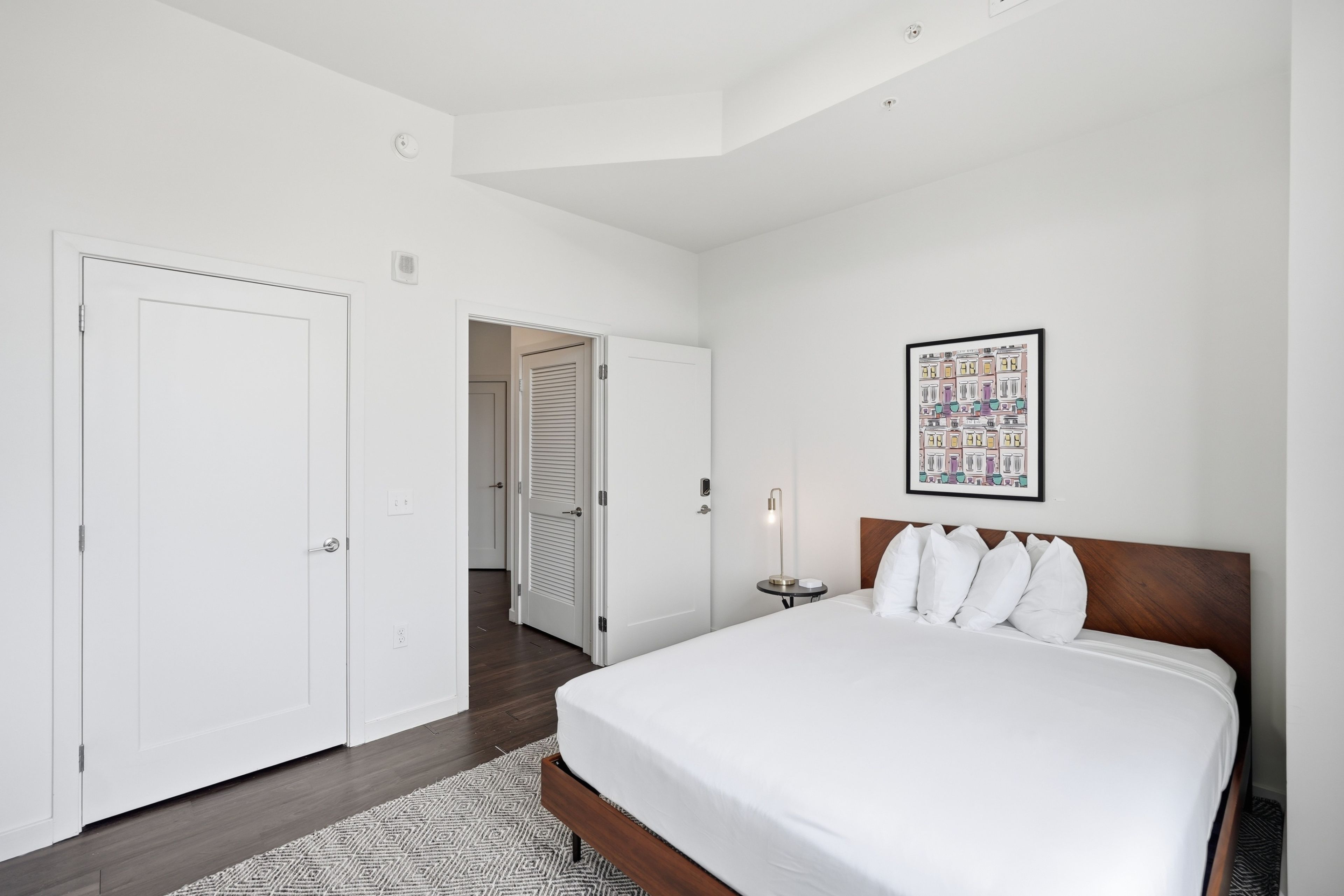 Una habitación de hotel con una cama grande, una mesita de noche, un cuadro en la pared y una puerta que da a otra habitación.