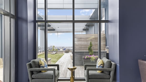 A modern room with large windows, two grey sofas, a small wooden table, and a view of a garden and building in the background. - Placemakr Marymount Ballston (Arlington)