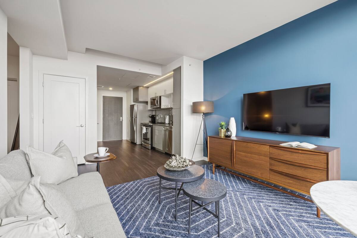 A modern living room with a sofa, a coffee table, a TV, and a kitchen in the background.
