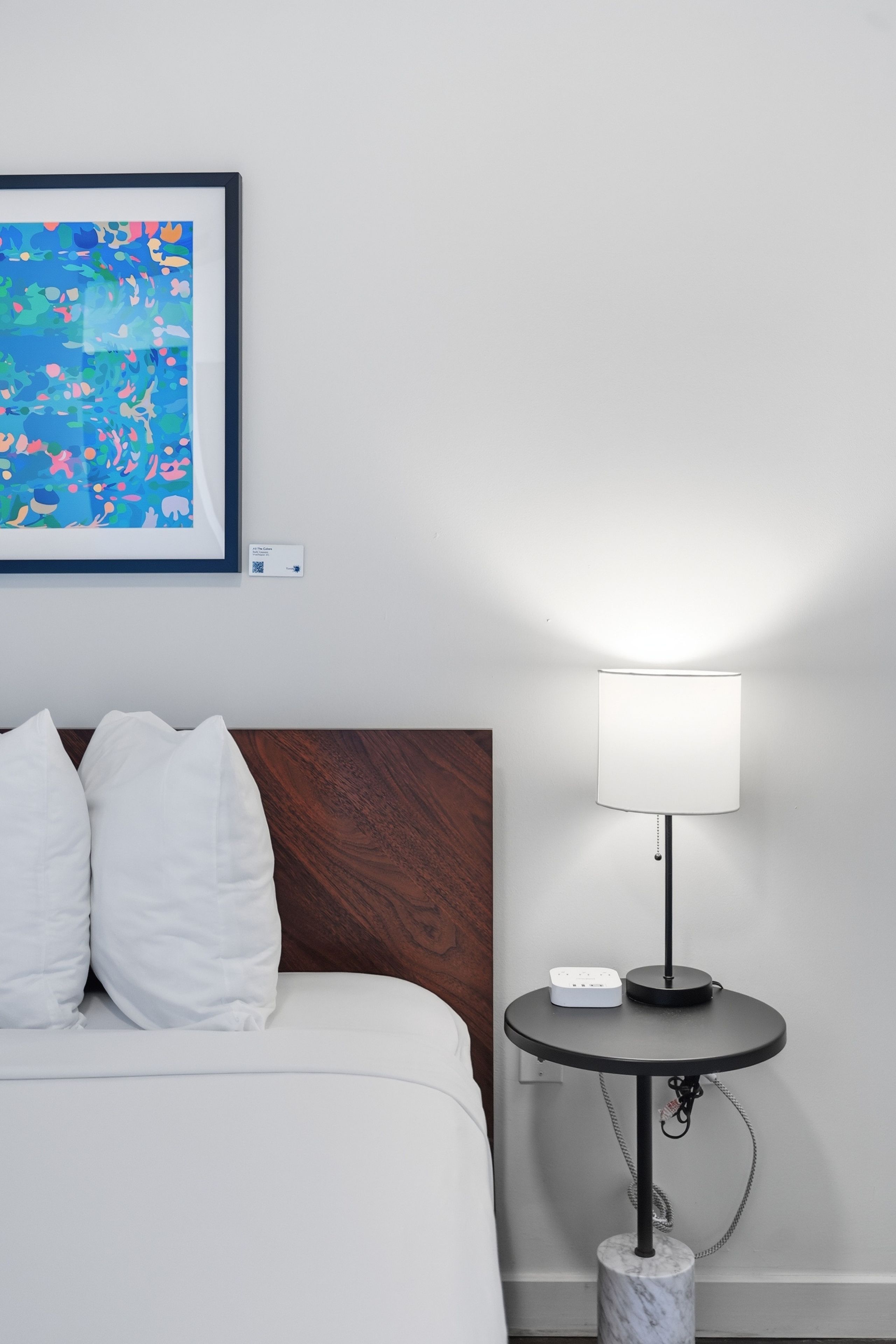 A neatly made bed with white bedding and pillows, a small round table with a lamp, and a framed abstract painting on the wall.
