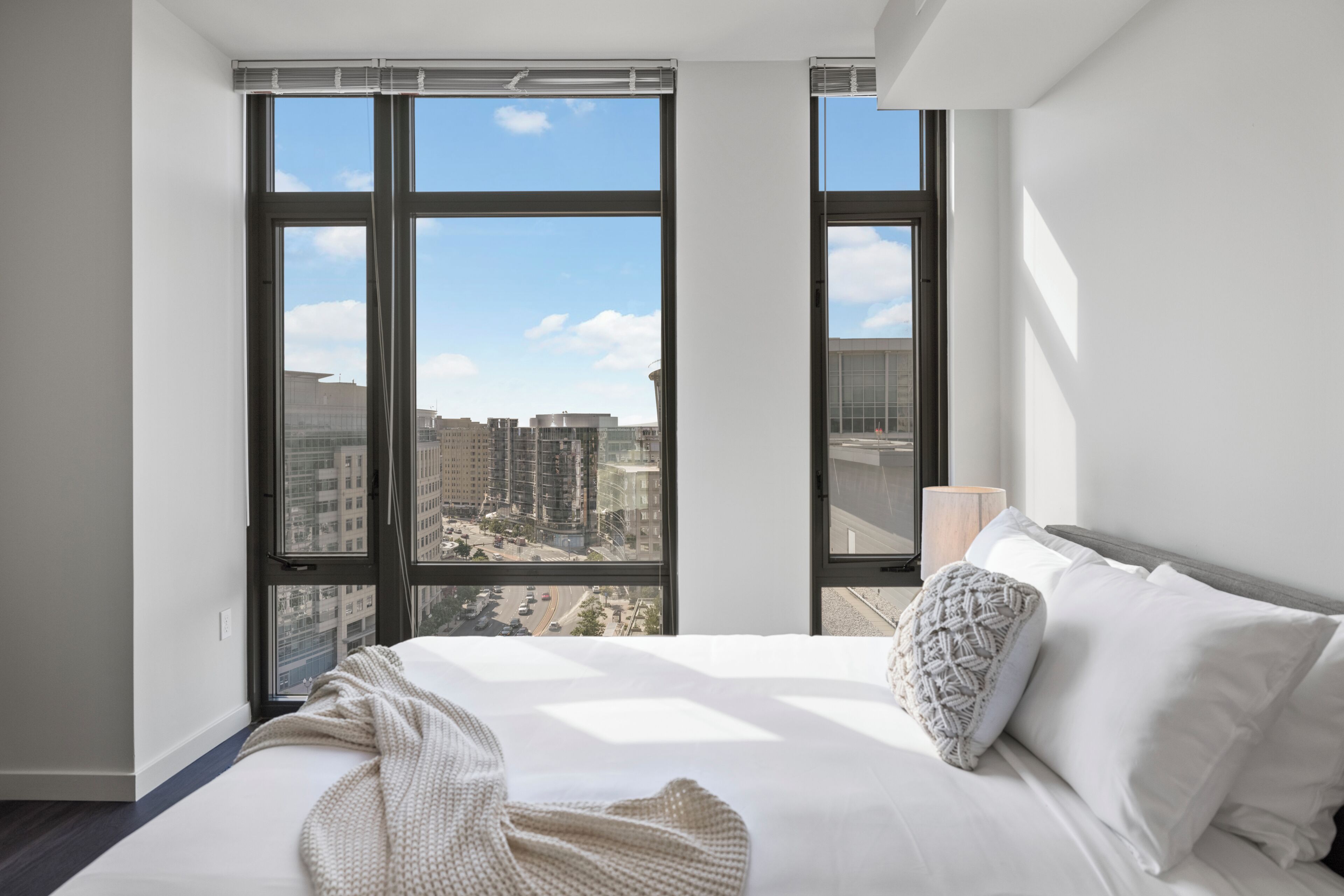 Una habitación de hotel moderna con una cama grande, ropa de cama blanca y vistas de la ciudad a través de ventanales de piso a techo.