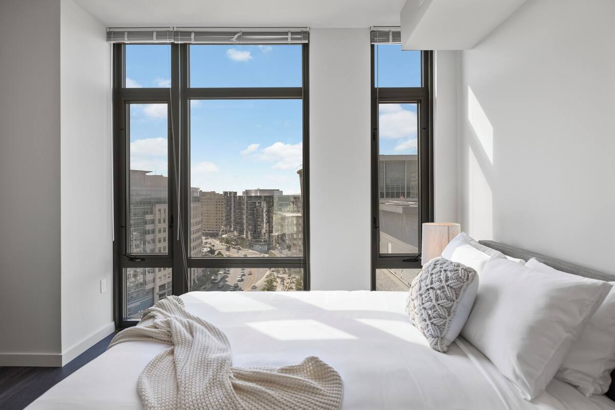 Quarto de hotel moderno com uma cama grande, roupa de cama branca e vista da cidade através de janelas do chão ao teto.