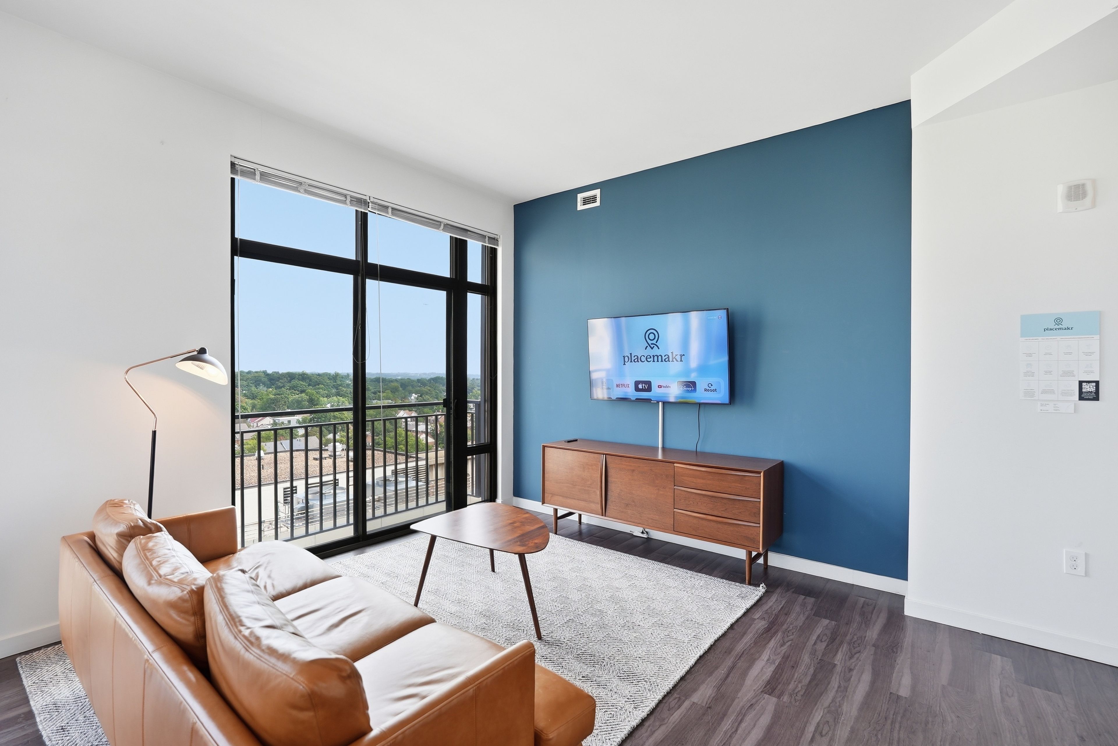 A modern living room with a leather sofa, a coffee table, a flat-screen TV mounted on a blue wall, and a view through large windows.