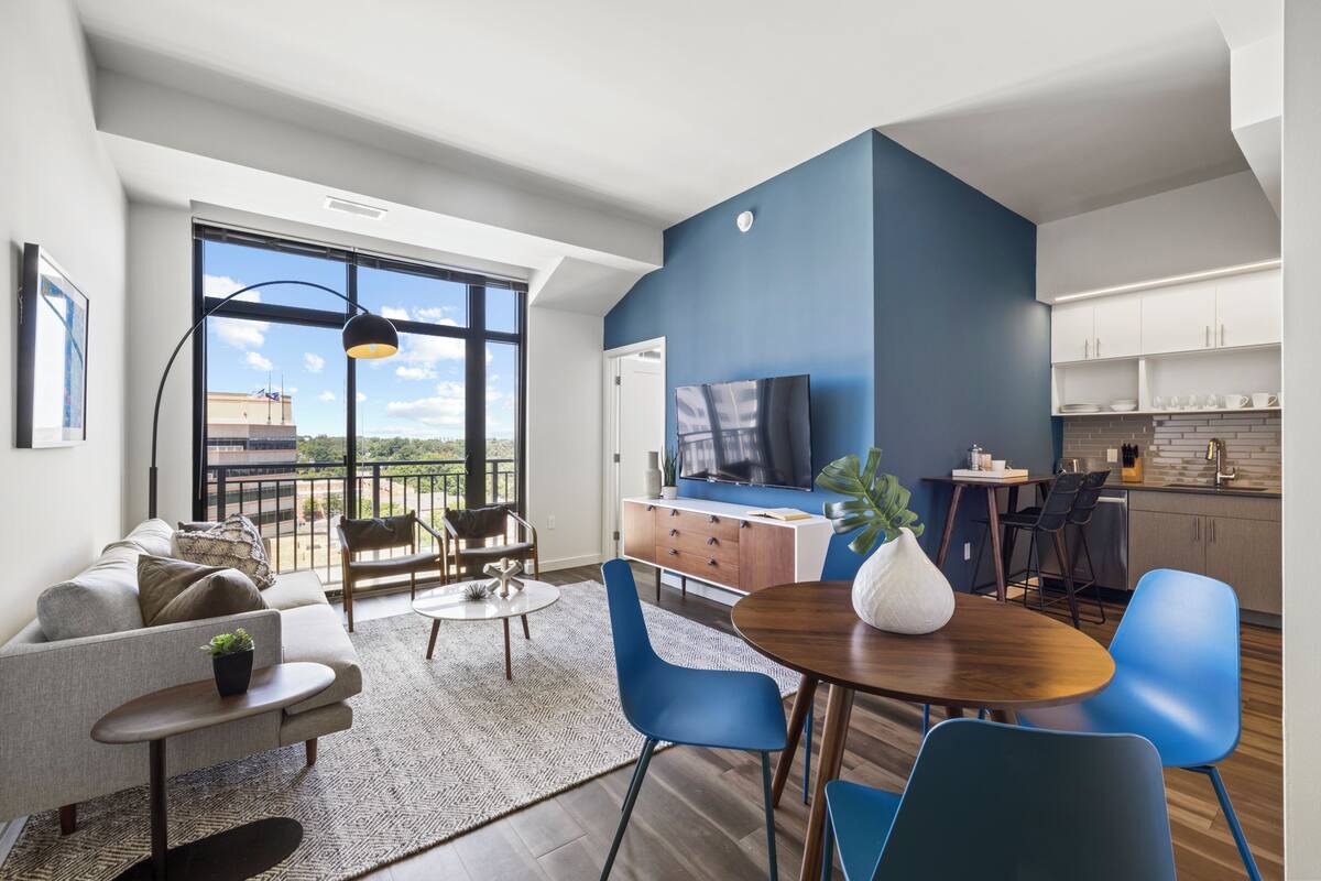 A modern living room with a sofa, coffee table, and blue chairs, a dining area with a wooden table and chairs, and a kitchen with white cabinets and a blue accent wall.