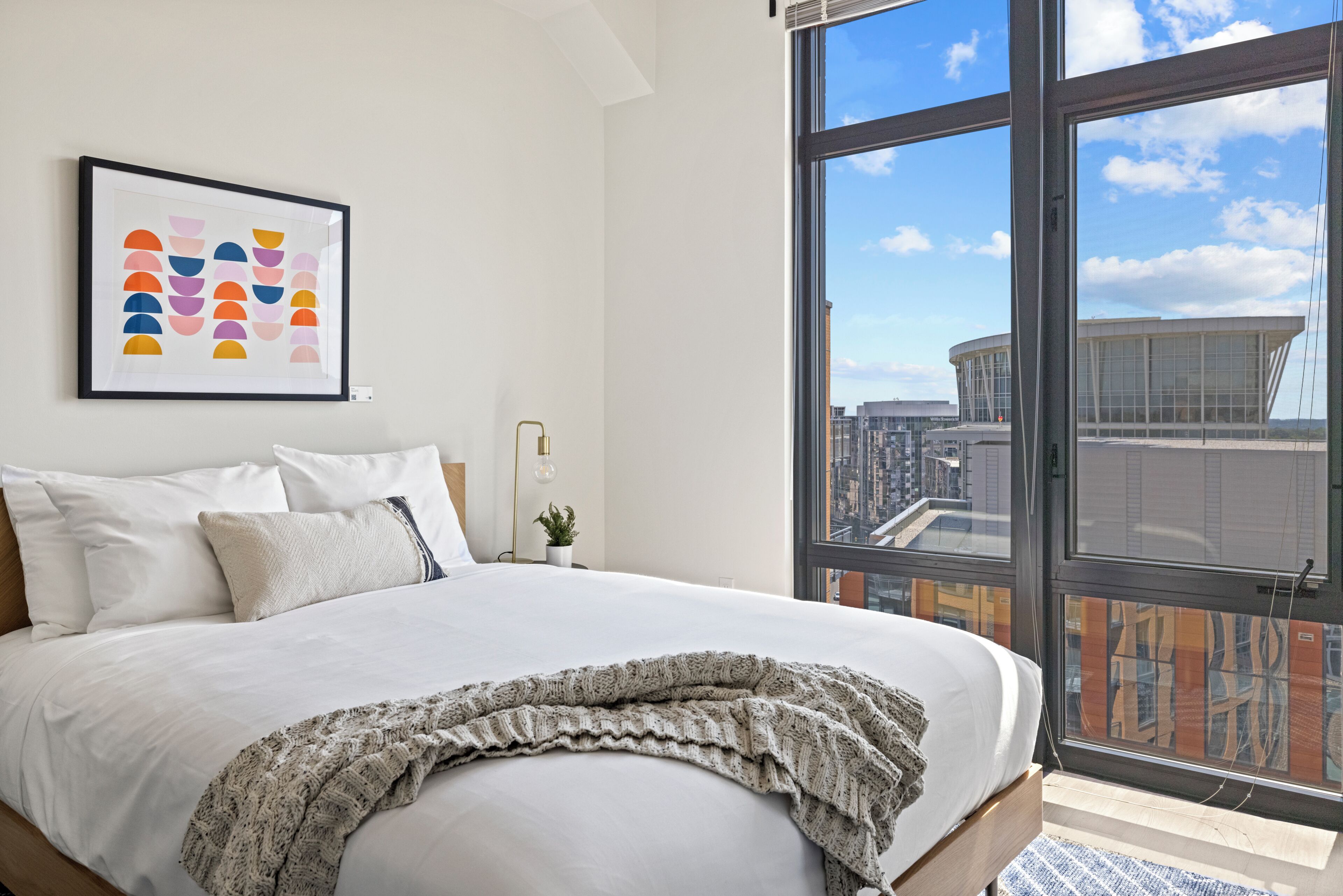 A modern bedroom with a large bed, a framed artwork, a potted plant, and a city view through large windows.