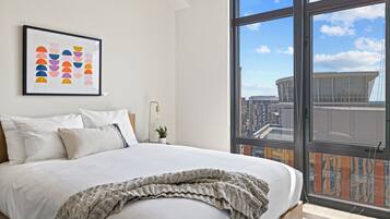 A modern bedroom with a large bed, a framed artwork, a potted plant, and a city view through large windows.