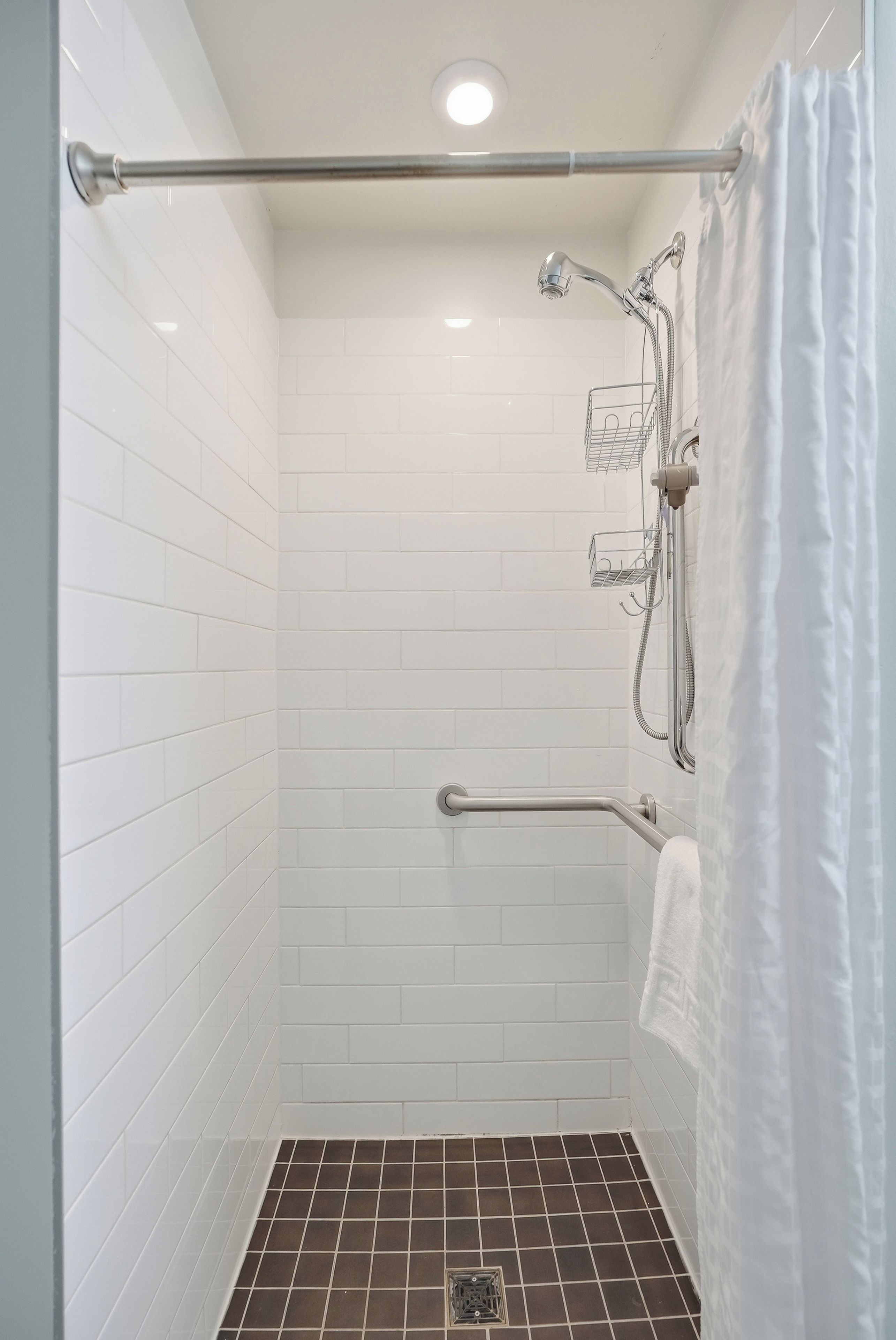 A shower area with white tiled walls, a showerhead, a handrail, and a white curtain.
