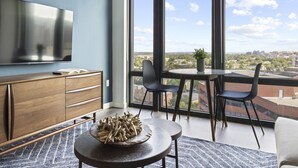 A modern living room with a large window, a flat-screen TV, a wooden cabinet, a round table with chairs, and a decorative bowl. - Placemakr Marymount Ballston (Arlington)