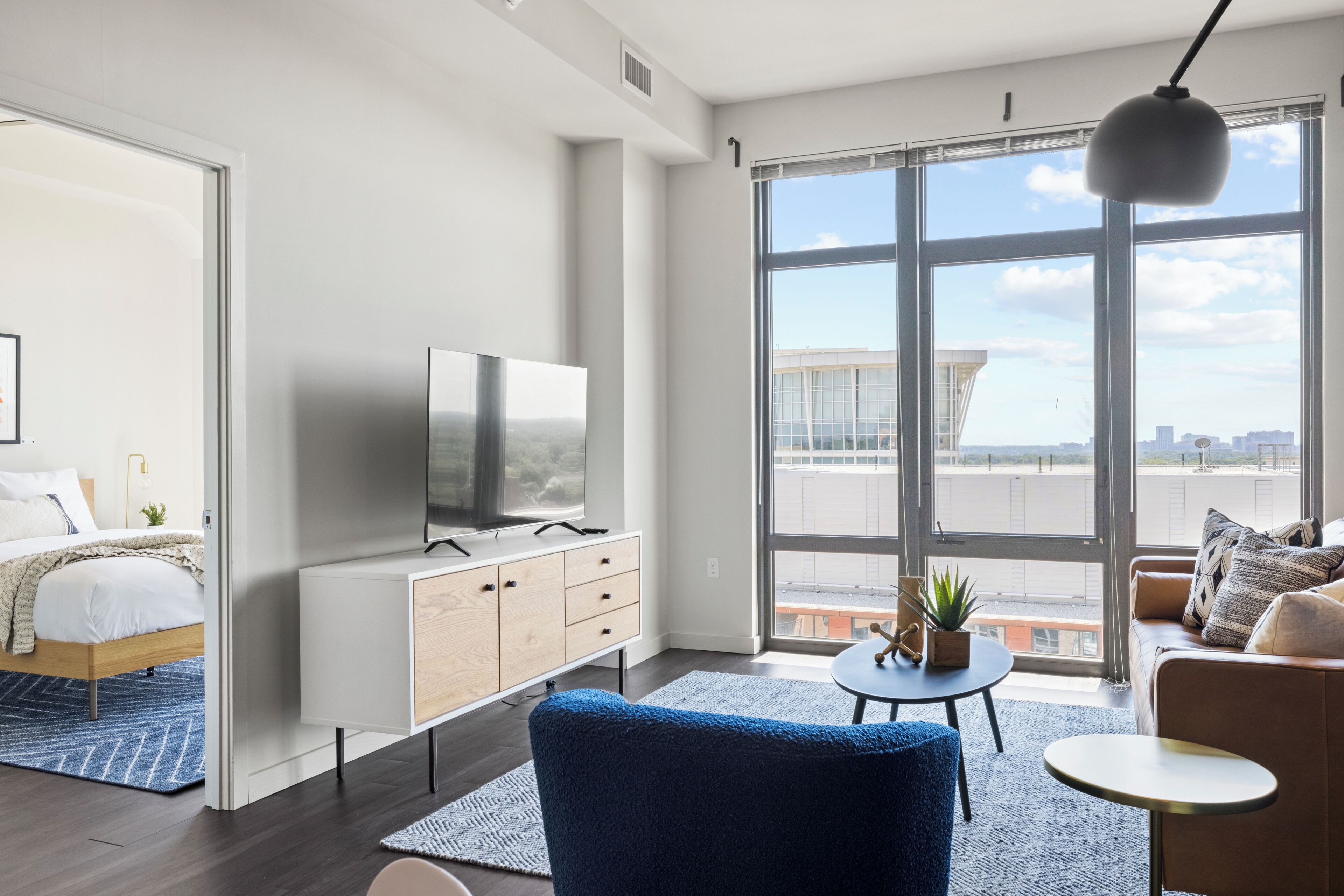 A modern hotel room with a large window, a bed, a TV, a sofa, and a small table.