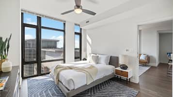 Une chambre moderne avec un grand lit, un ventilateur de plafond et une vue sur la ville grâce à de grandes fenêtres du sol au plafond.