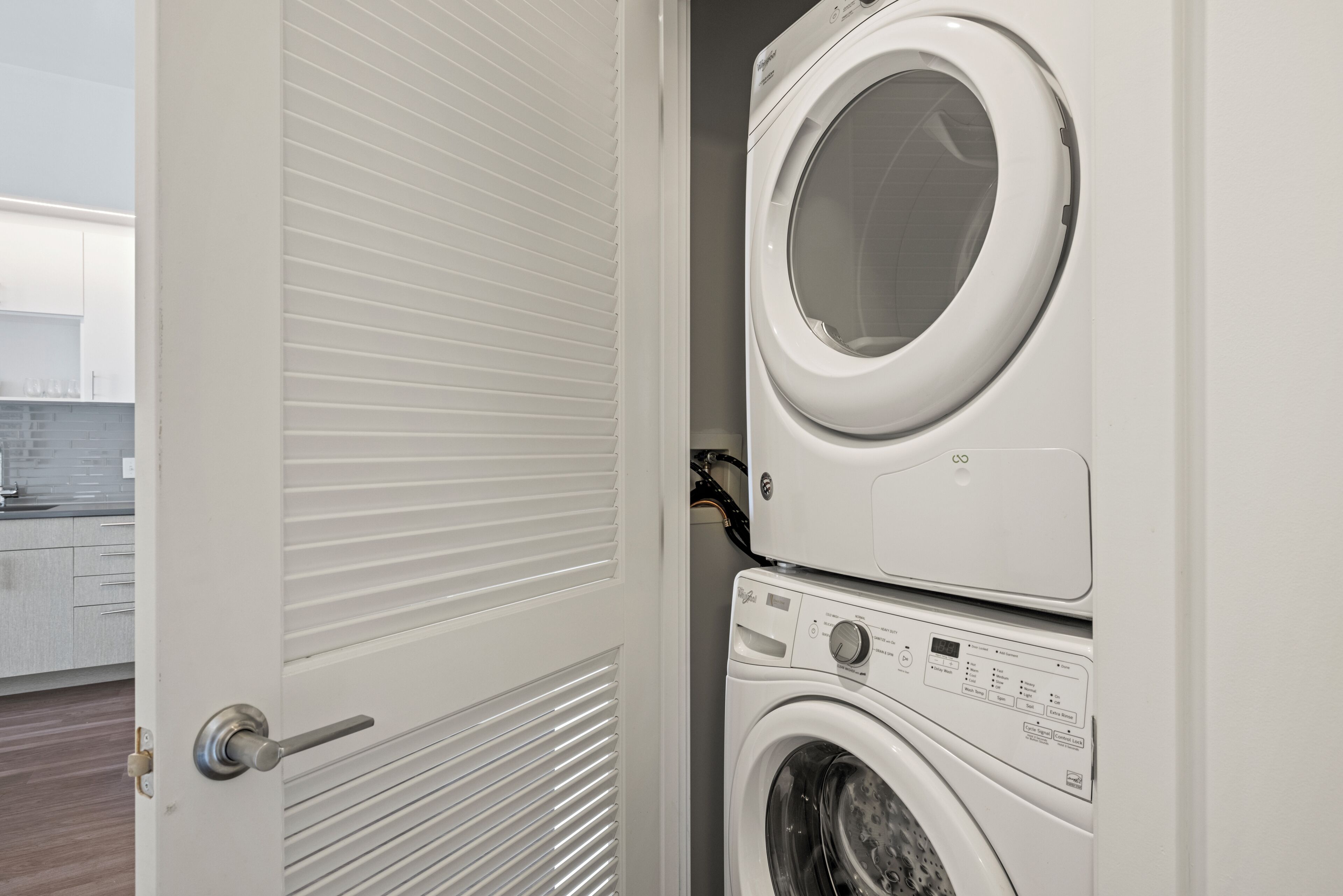 A laundry room with a stacked washer and dryer, a door with a handle, and a glimpse of a kitchen area in the background.