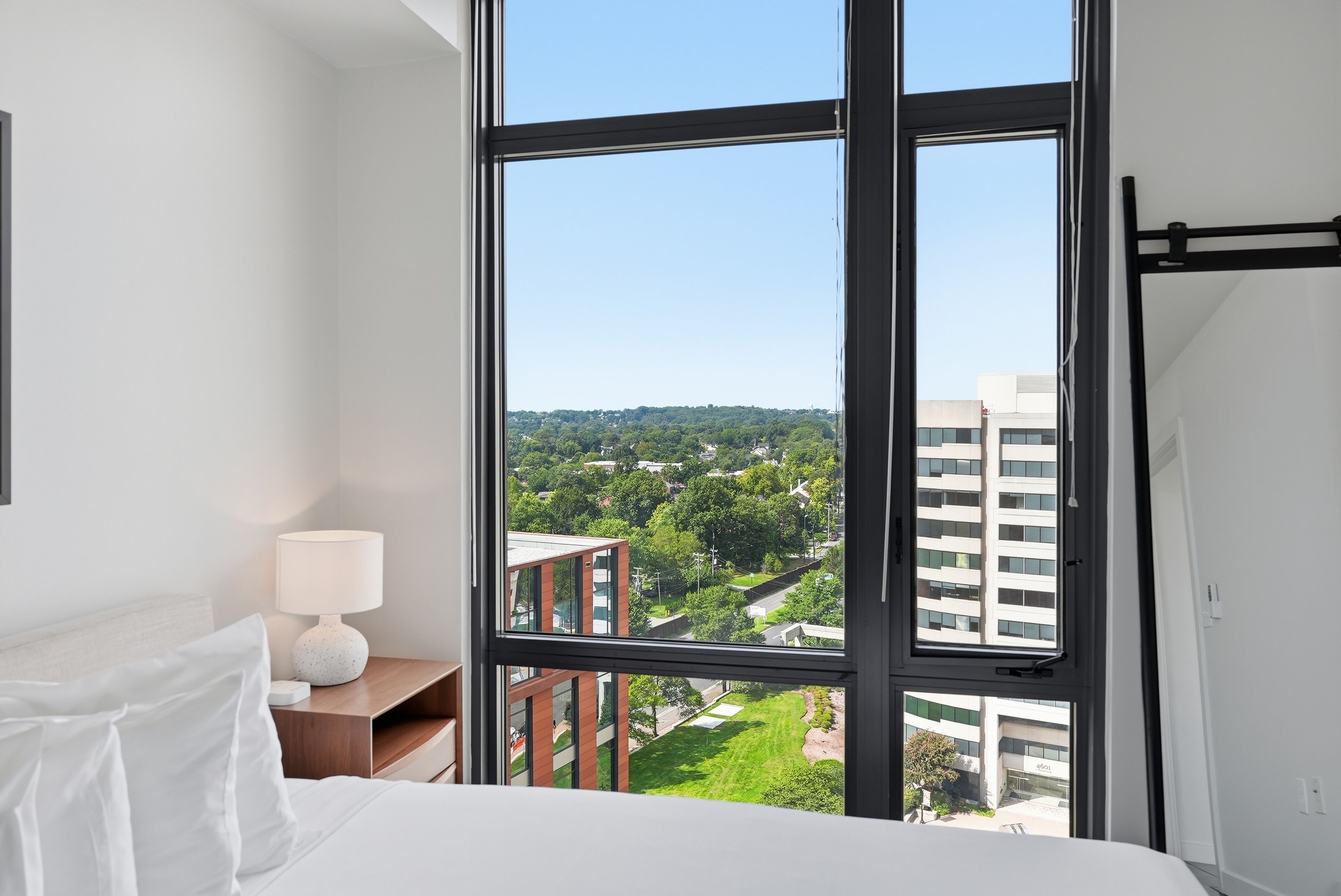 A hotel room with a large window, a bed, a bedside table with a lamp, and a view of the cityscape.