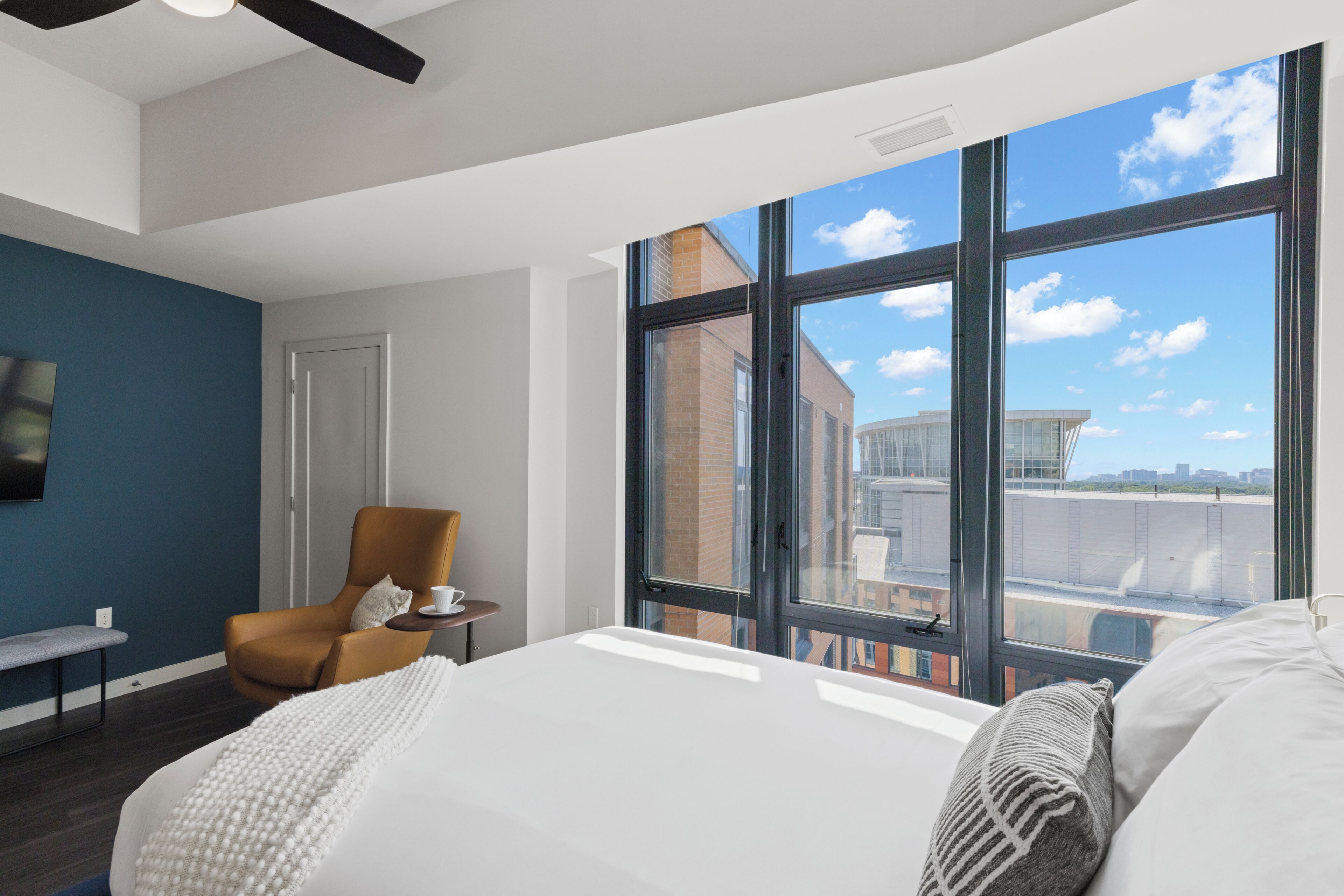 Une chambre d’hôtel moderne dotée d’un grand lit, d’un coin salon avec un fauteuil et une table, et d’une grande fenêtre donnant sur la ville.