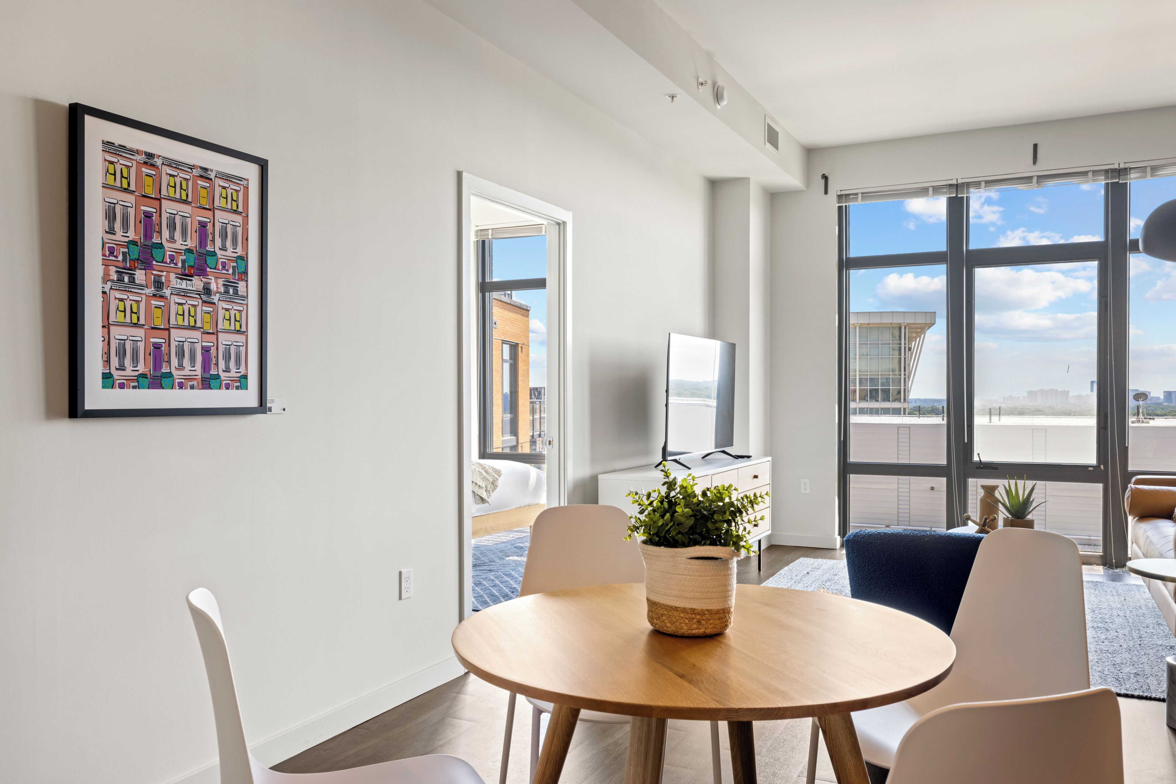 A modern living room with a round wooden table, white chairs, a large window with a city view, and a framed artwork on the wall.