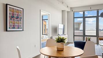 A modern living room with a round wooden table, white chairs, a large window with a city view, and a framed artwork on the wall.