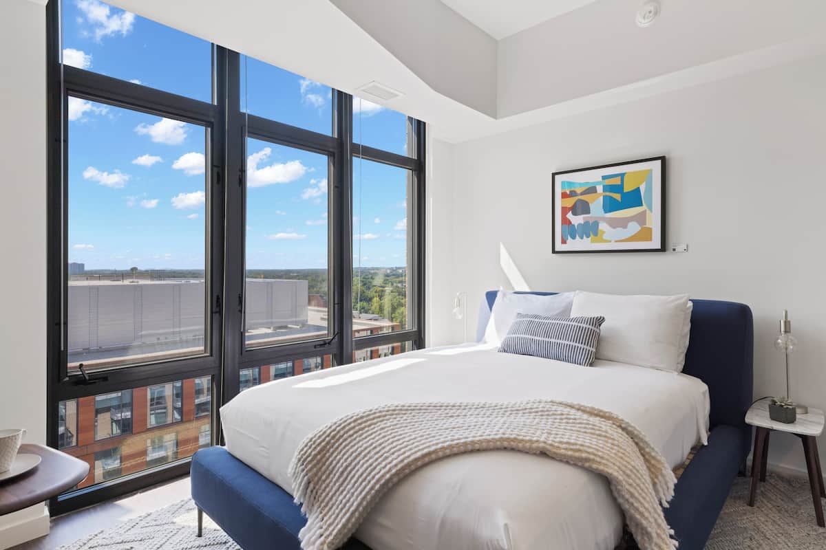 A modern bedroom with a large bed, a view through floor-to-ceiling windows, and a piece of abstract art on the wall.