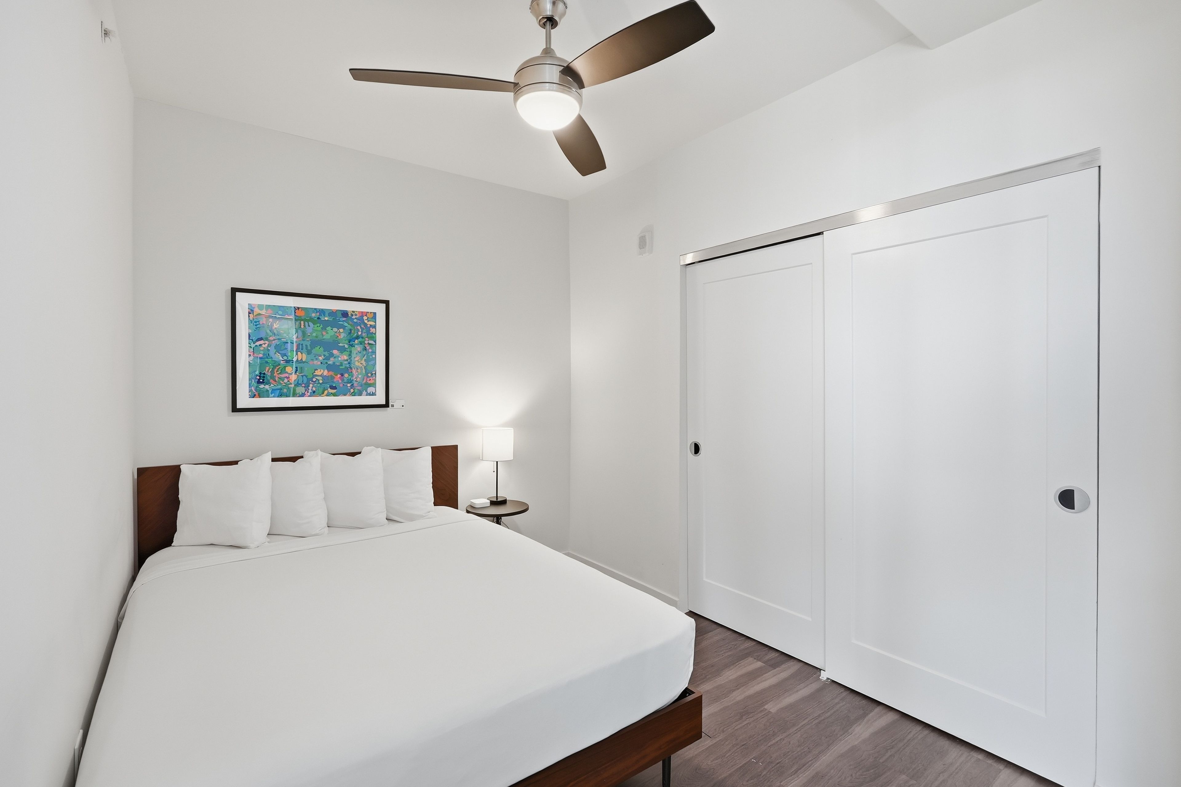 Une chambre d’hôtel avec un lit, un ventilateur de plafond, une table de chevet avec une lampe, un tableau encadré au mur et une armoire blanche.