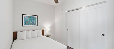 Une chambre d’hôtel avec un lit, un ventilateur de plafond, une table de chevet avec une lampe, un tableau encadré au mur et une armoire blanche.