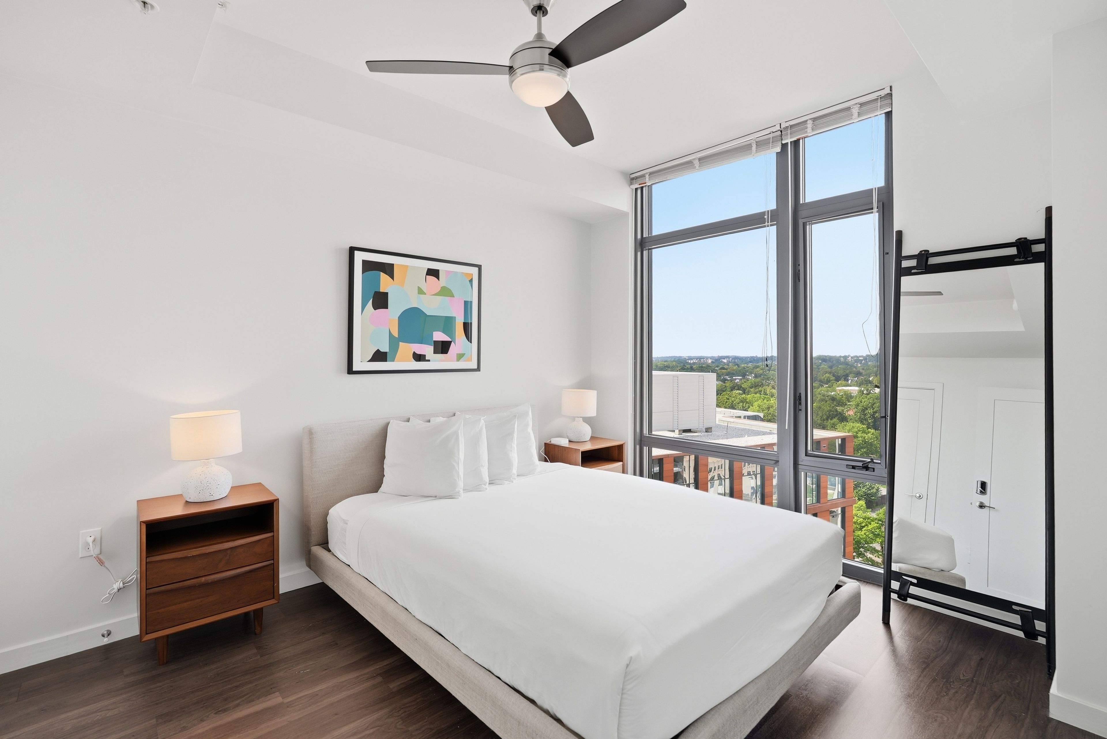 A modern bedroom with a large bed, two bedside tables, a ceiling fan, a window with a view, and a framed abstract painting.