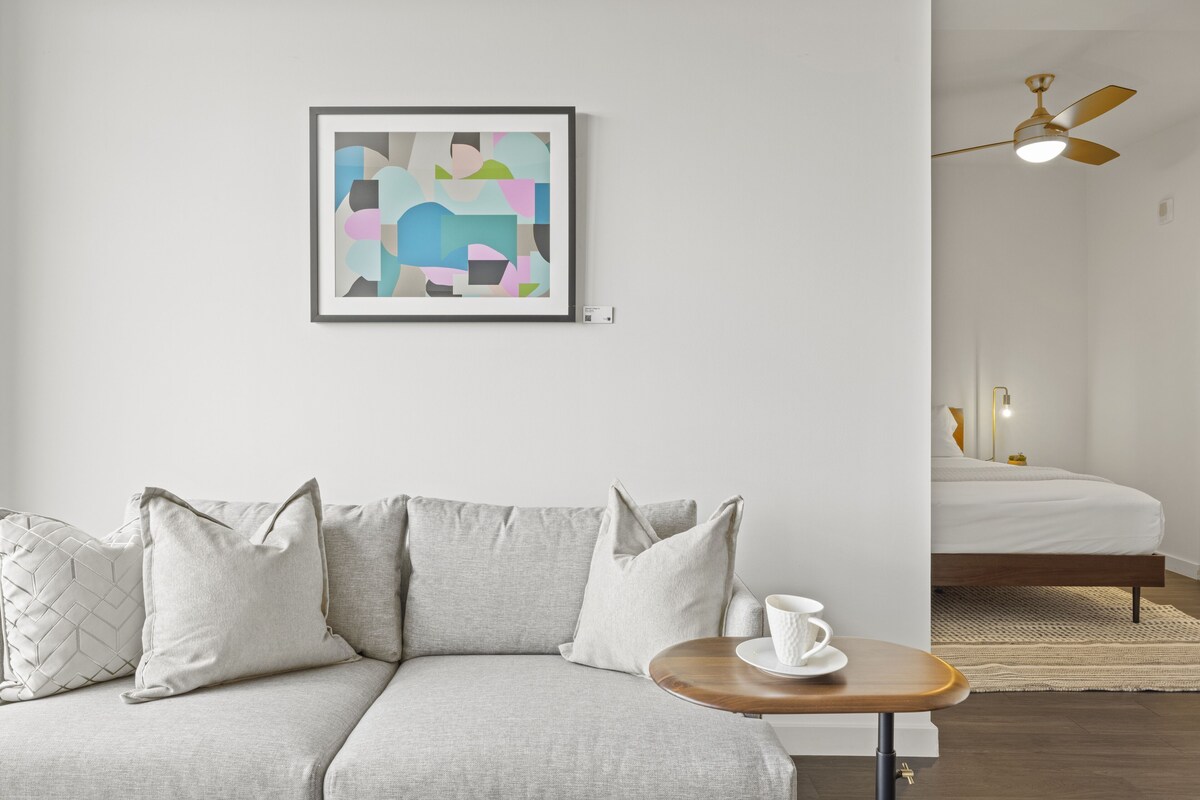 A modern room with a grey sofa, a framed abstract painting, a wooden side table with a white cup, and a bed with a white duvet.