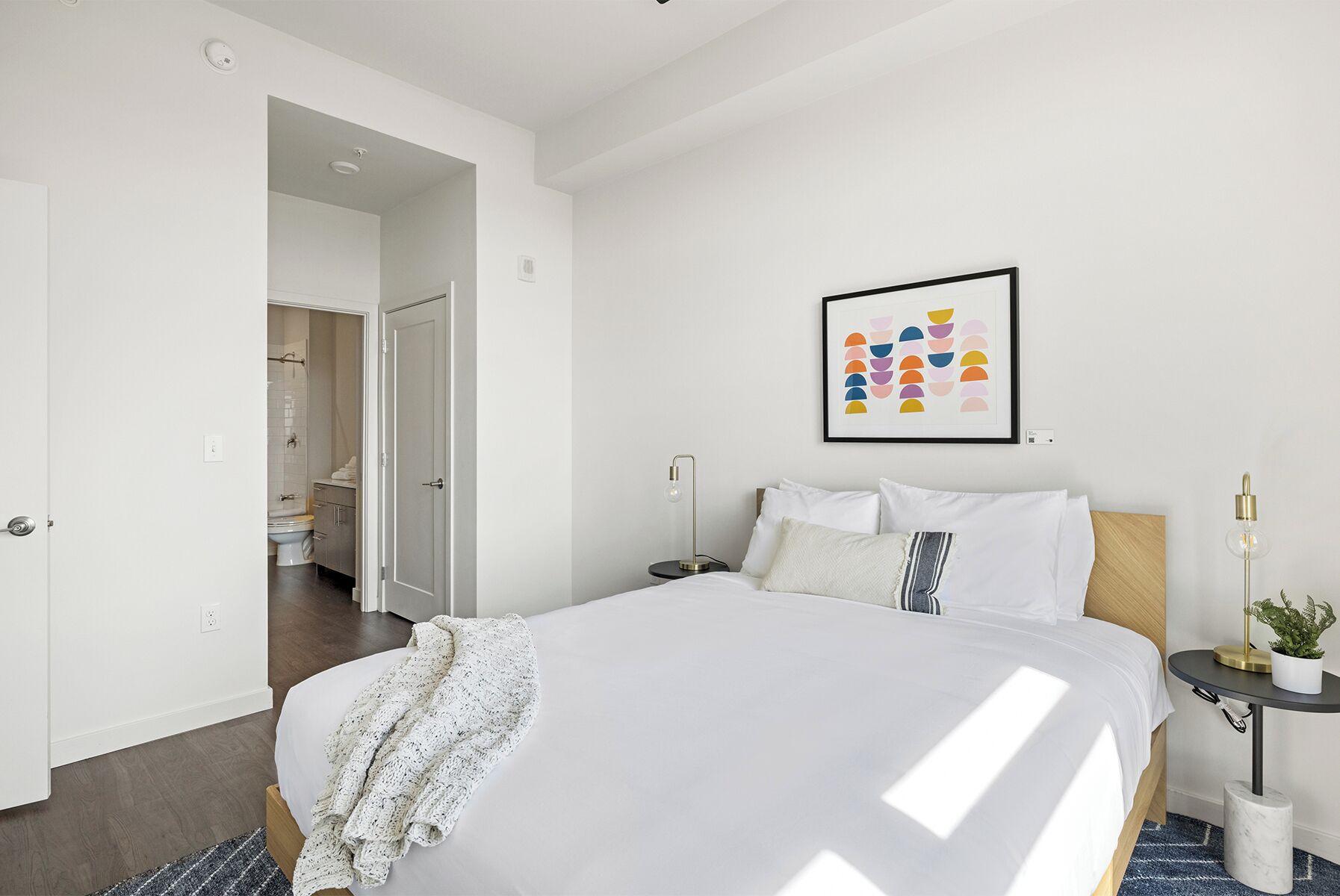 Habitación de hotel con una cama grande, una mesita de noche con lámpara, una obra de arte enmarcada en la pared y vista al baño.