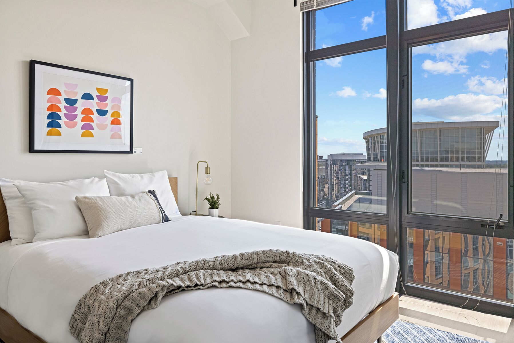 A modern hotel room with a large bed, a framed abstract art piece, a city view through a large window, and a small potted plant.