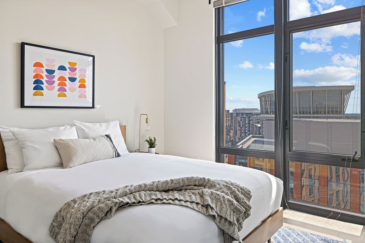 A modern hotel room with a large bed, a framed abstract art piece, a city view through a large window, and a small potted plant.