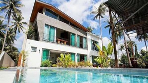 Un hotel moderno de dos pisos con piscina, rodeado de palmeras y plantas tropicales.
