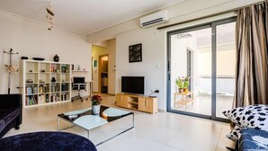Ein modernes Wohnzimmer mit Sofa, Couchtisch, Fernseher, Bücherregal und einer Glastür zum Balkon.