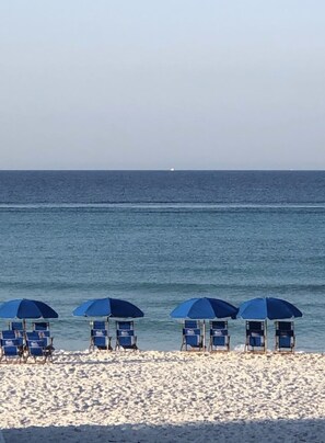 Ein Strand mit blauen Liegestühlen und Sonnenschirmen, einem sandigen Ufer und einem ruhigen Meer im Hintergrund.