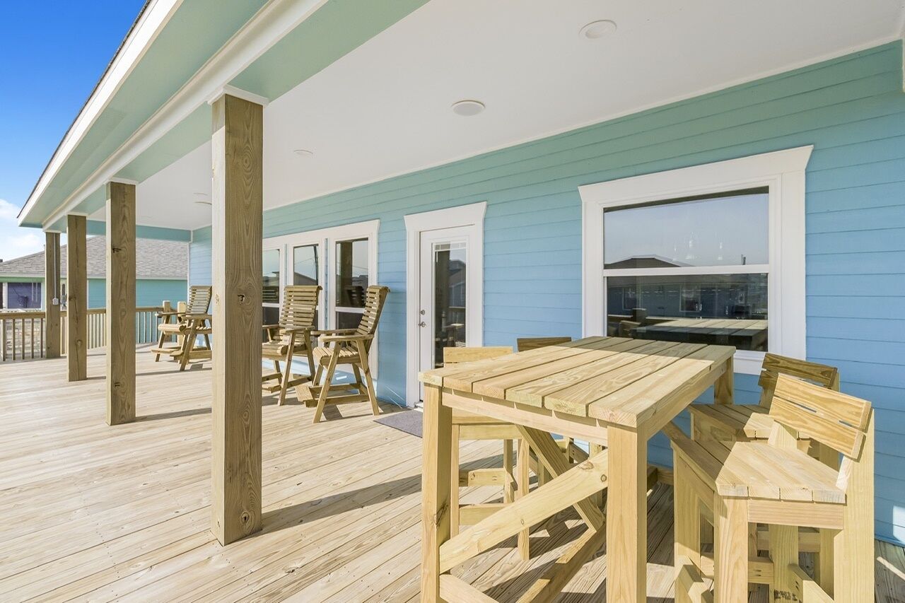 A wooden deck with a table and chairs, a covered area with a view of the ocean, and a building with blue siding.