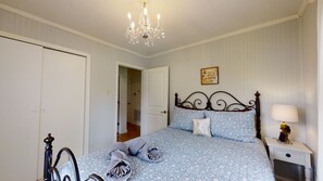 Een slaapkamer met een bed, nachtkastje, lamp, een kroonluchter en een deur naar een andere kamer.